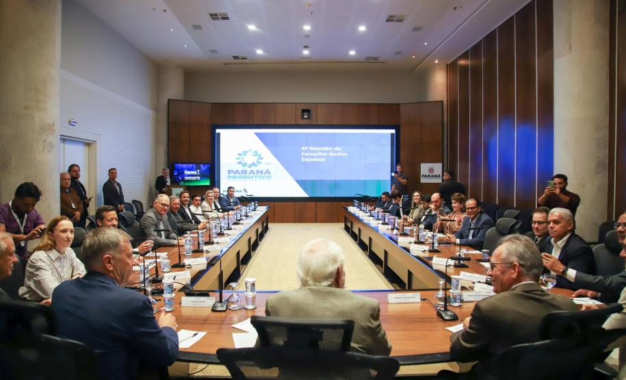Reunião presencial do Conselho Gestor Estadual em auditório com grande mesa em formato de U. Mais de 20 participantes, vestindo trajes sociais e casuais finos, estão sentados ao redor da mesa. Ao fundo, um painel de LED exibe o slide da "4ª Reunião do Conselho Gestor Estadual", com o logotipo "Paraná Produtivo".