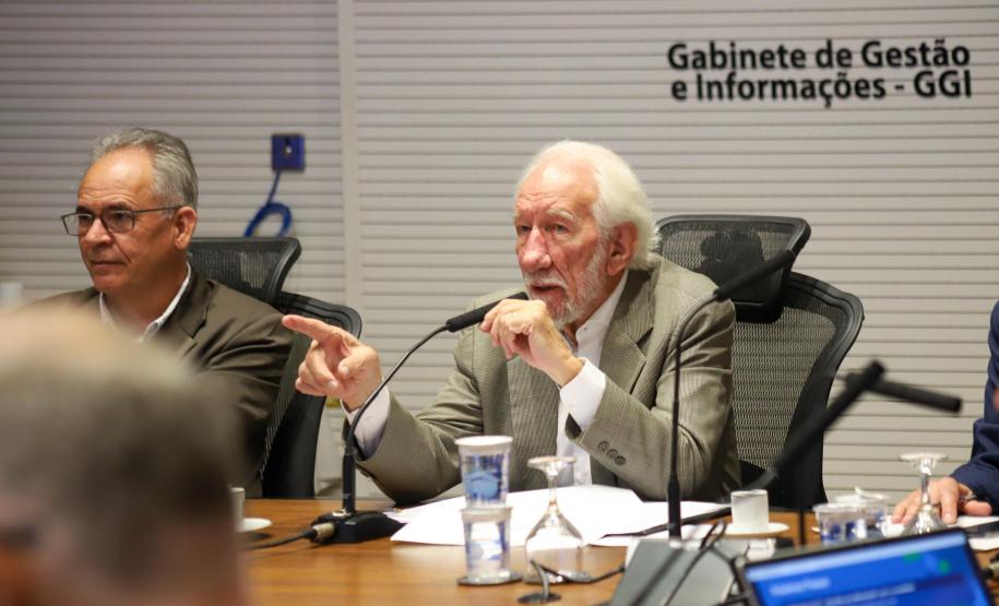 Fotografia foca em Darci Piana, Vice-Governador, de terno bege e cabelos brancos, falando ao microfone enquanto aponta com o dedo indicador da mão direita. Ele está sentado à mesa de reunião, olhando para a esquerda da imagem. À sua esquerda, sentado ao seu lado, está Domingos Trevisan Filho, diretor-geral da SEPL, de óculos e terno marrom, ouvindo atentamente. Sobre a mesa, há copos d'água e microfones. Ao fundo, uma parede branca exibe o texto: 'Gabinete de Gestão e Informações - GGI'.