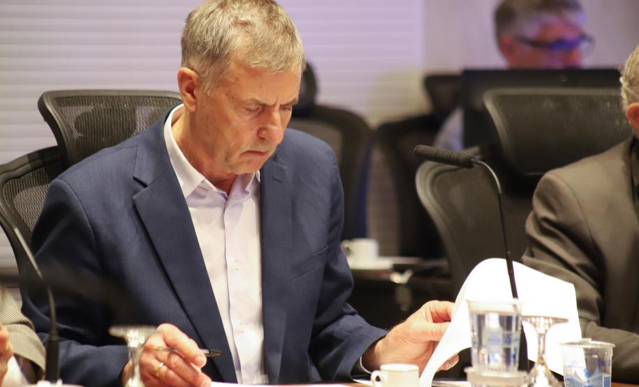 Fotografia em plano médio foca em Luizão Goulart, secretário da Administração e da Previdência, sentado à mesa de reunião. Ele está com a cabeça levemente abaixada, focado na leitura de documentos. Ele veste paletó azul-marinho sobre camisa social clara. Segura uma caneta na mão direita sobre um papel e manuseia outras folhas com a mão esquerda. À sua frente, copos d'água e uma xícara de café. Outros participantes desfocados estão ao seu redor.