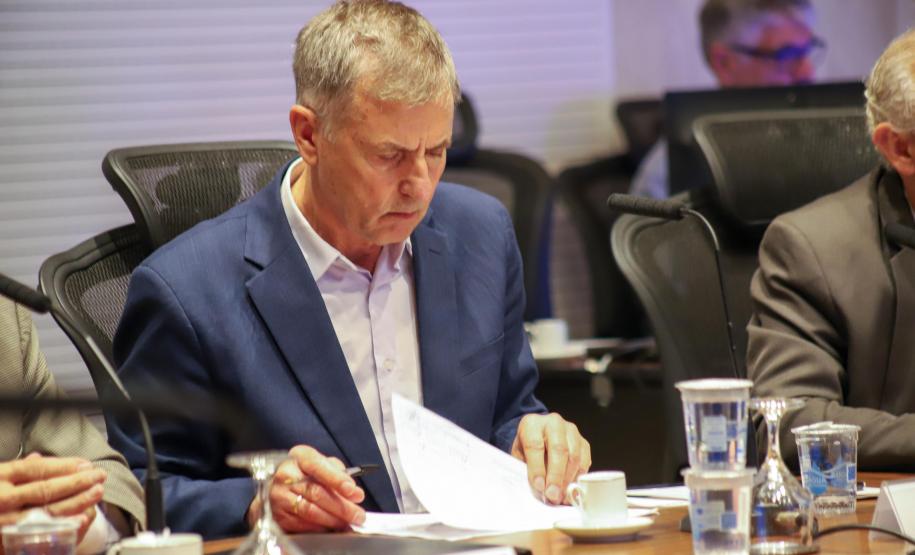 Fotografia em plano médio foca em Luizão Goulart, secretário da Administração e da Previdência, sentado à mesa de reunião. Ele está com os cabelos grisalhos abaixados, lendo atentamente documentos impressos sobre a mesa. Veste paletó azul-marinho e camisa social clara. Segura uma caneta na mão direita e folheia o papel com a esquerda. À sua frente, copos d'água de vidro e plástico, e uma xícara de café vazia. Ao fundo, desfocadas, aparecem outras pessoas sentadas à mesa.
