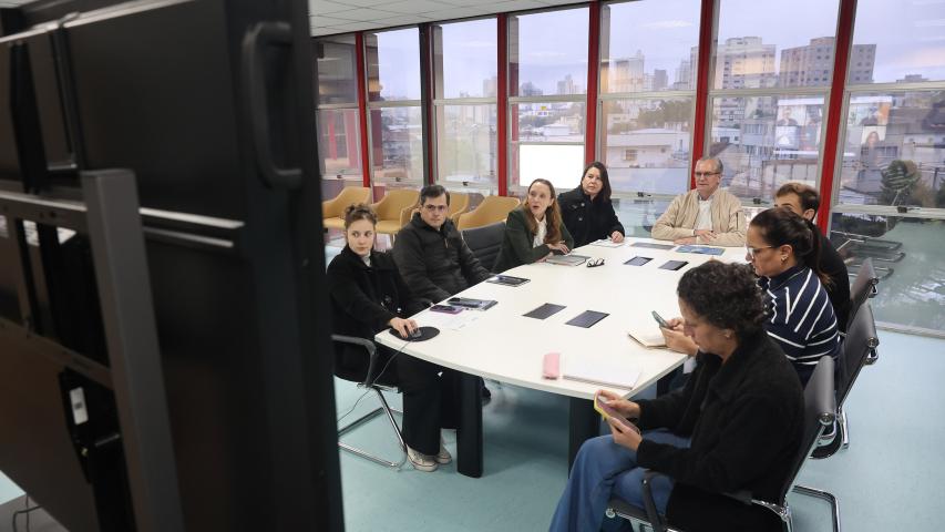 Pessoas sentadas ao redor de uma grande mesa branca em uma sala de reuniões envidraçada, participando de uma videoconferência exibida em um grande monitor em primeiro plano.